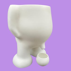 May include: A white ceramic planter with a unique, abstract design. The planter has a large, rounded body supported by three bulbous legs. A small, matching cup sits beside it. The background is a solid lavender color.