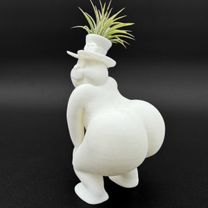 May include: White ceramic planter shaped like a person wearing a hat, with a small green plant growing out of the top of the hat.
