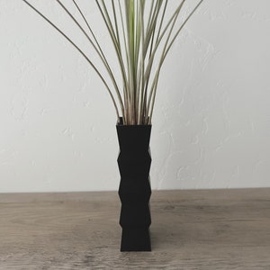 May include: A black vase with a geometric pattern holds a bunch of green and white plants.