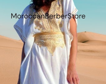 Puede incluir: Una kaftán blanca con bordados dorados y un cinturón dorado. La kaftán la lleva puesta una persona de pie en un paisaje desértico. El texto "MoroccanBerberStore" es visible en la imagen.