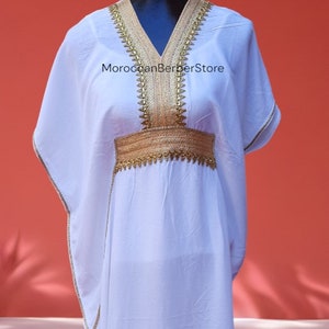 May include: White kaftan with gold embroidery around the neckline and waist. The kaftan has a V-neck and long sleeves. The text "MoroccanBerberStore" is visible on the front of the garment.