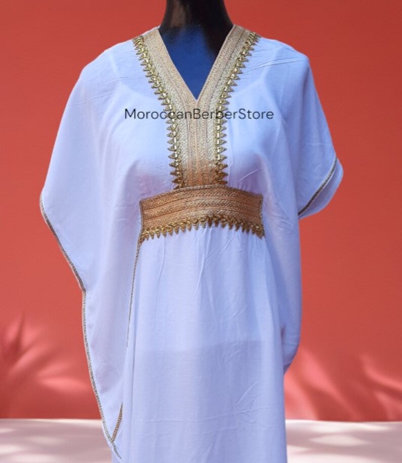 May include: White kaftan with gold embroidery around the neckline and waist. The kaftan has a V-neck and long sleeves. The text "MoroccanBerberStore" is visible on the front of the garment.