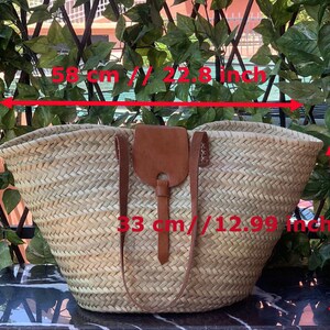 May include: Woven straw tote bag with brown leather accents. The bag measures 22.8 inches wide and 12.99 inches high. Features a leather flap closure and straps. Ideal for carrying items.