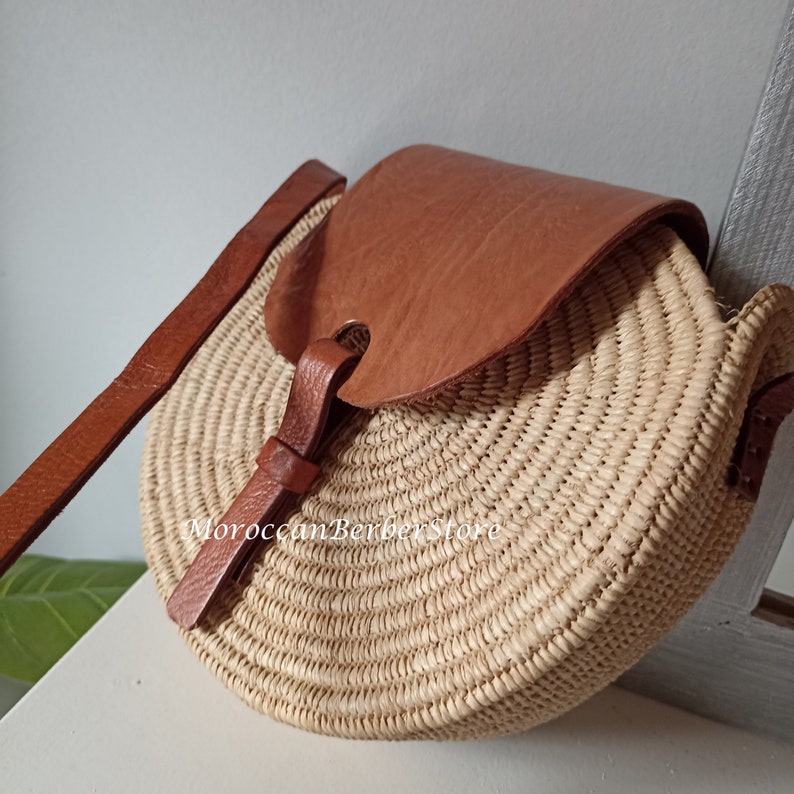 May include: A round straw bag with a brown leather flap and strap. The bag has a woven design and is perfect for carrying everyday essentials.