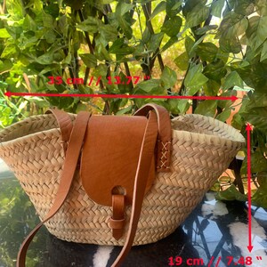 May include: Woven straw tote bag with a brown leather flap and strap. The bag is 13.77 inches wide and 7.48 inches tall. The bag has a natural color and is suitable for carrying items.