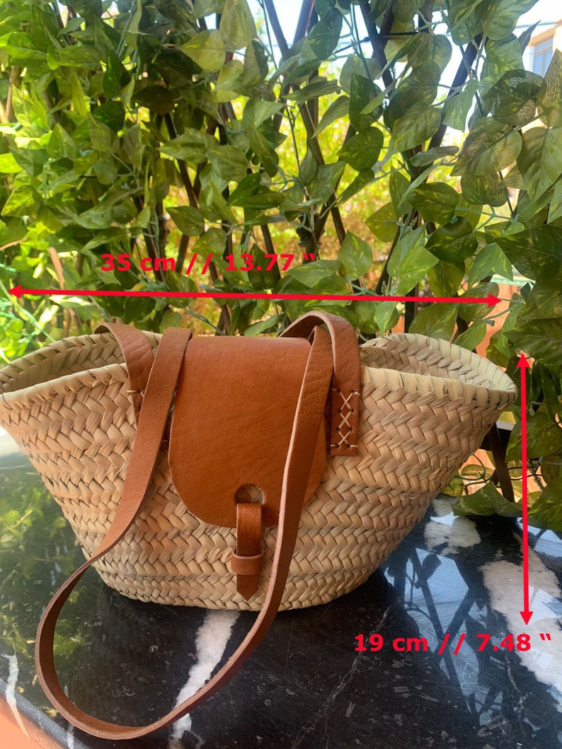 May include: Woven straw tote bag with a brown leather flap and strap. The bag is 13.77 inches wide and 7.48 inches tall. The bag has a natural color and is suitable for carrying items.