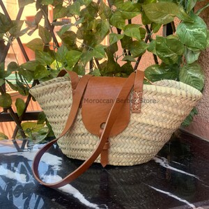 May include: A woven straw tote bag with a brown leather flap and shoulder strap. The bag has a rectangular shape with a rounded top. The text "Moroccan Berber Store" is visible on the bag.