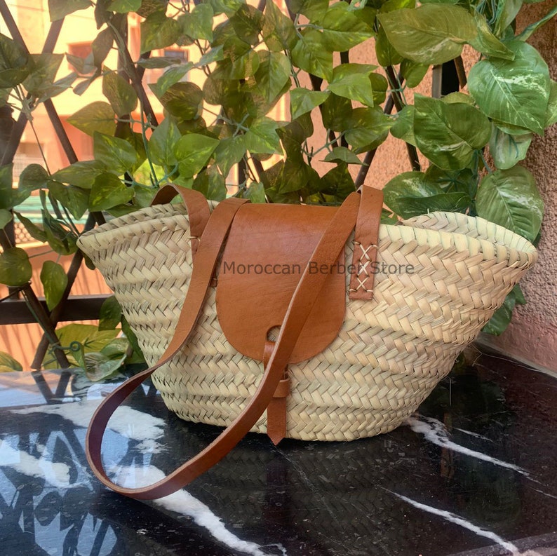 May include: A woven straw tote bag with a brown leather flap and shoulder strap. The bag has a rectangular shape with a rounded top. The text "Moroccan Berber Store" is visible on the bag.