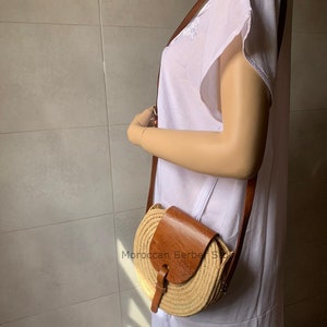 May include: A brown leather and straw crossbody bag with a flap closure and a long brown leather strap. The bag is worn by a mannequin wearing a white dress. The text "Moroccan Berber Style" is visible on the bag.