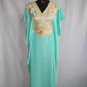 Puede incluir: Una kaftan verde menta con bordados dorados. La kaftan tiene un escote en V y mangas cortas. El bordado es intrincado y detallado, y cubre el escote, los hombros y el pecho de la prenda.