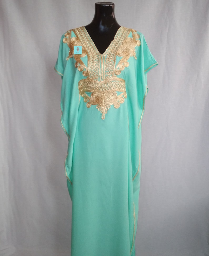 Puede incluir: Una kaftan verde menta con bordados dorados. La kaftan tiene un escote en V y mangas cortas. El bordado es intrincado y detallado, y cubre el escote, los hombros y el pecho de la prenda.