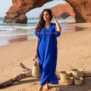 May include: A vibrant royal blue kaftan dress featuring a deep V-neckline with intricate tonal embroidery around the chest area. The garment has short, loose sleeves and a long, flowing silhouette that reaches the ankles, made of lightweight fabric.