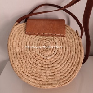 May include: A round straw bag with a brown leather flap and a long brown leather strap. The bag has a woven design and the text "Moroccan Berber Store" is visible on the front.