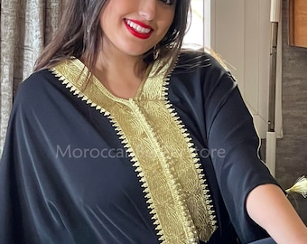 May include: A black kaftan with gold embroidery. The kaftan has a V-neck and long sleeves. The embroidery is intricate and detailed, and it covers the neckline and the front of the garment. The kaftan is worn by a person with long dark hair and red lipstick.