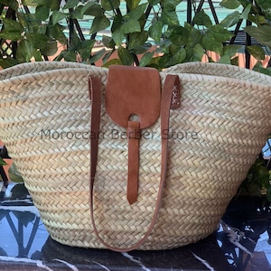 May include: Woven straw tote bag with brown leather accents. The bag features a large, open design with sturdy handles and a leather flap closure. The text "Moroccan Berber Store" is visible on the bag. This bag is perfect for carrying groceries or other items.