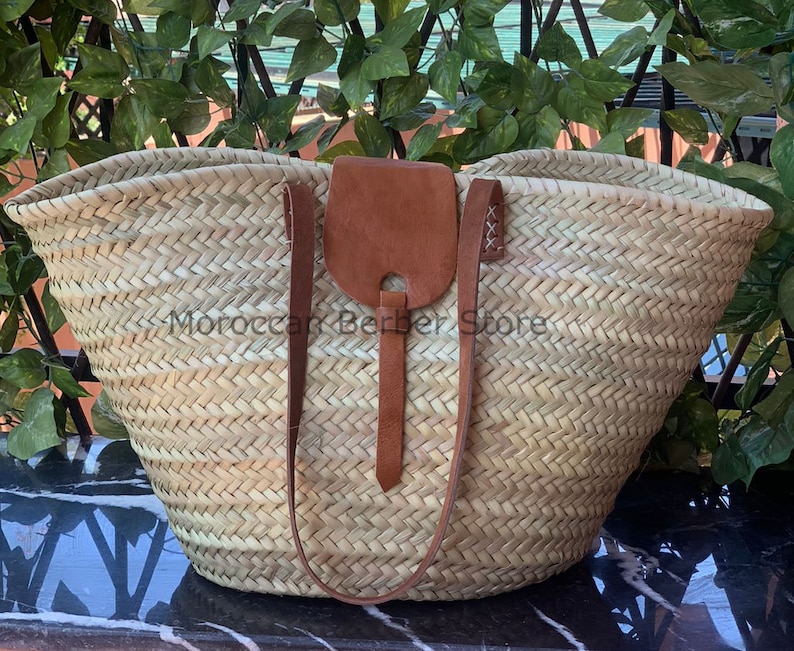 May include: Woven straw tote bag with brown leather accents. The bag features a large, open design with sturdy handles and a leather flap closure. The text "Moroccan Berber Store" is visible on the bag. This bag is perfect for carrying groceries or other items.