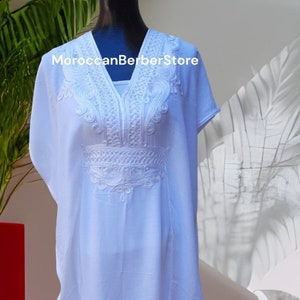 May include: A light blue kaftan with white embroidery. The embroidery features a geometric pattern around the neckline and a floral design on the chest. The text "MoroccanBerberStore" is visible on the garment.