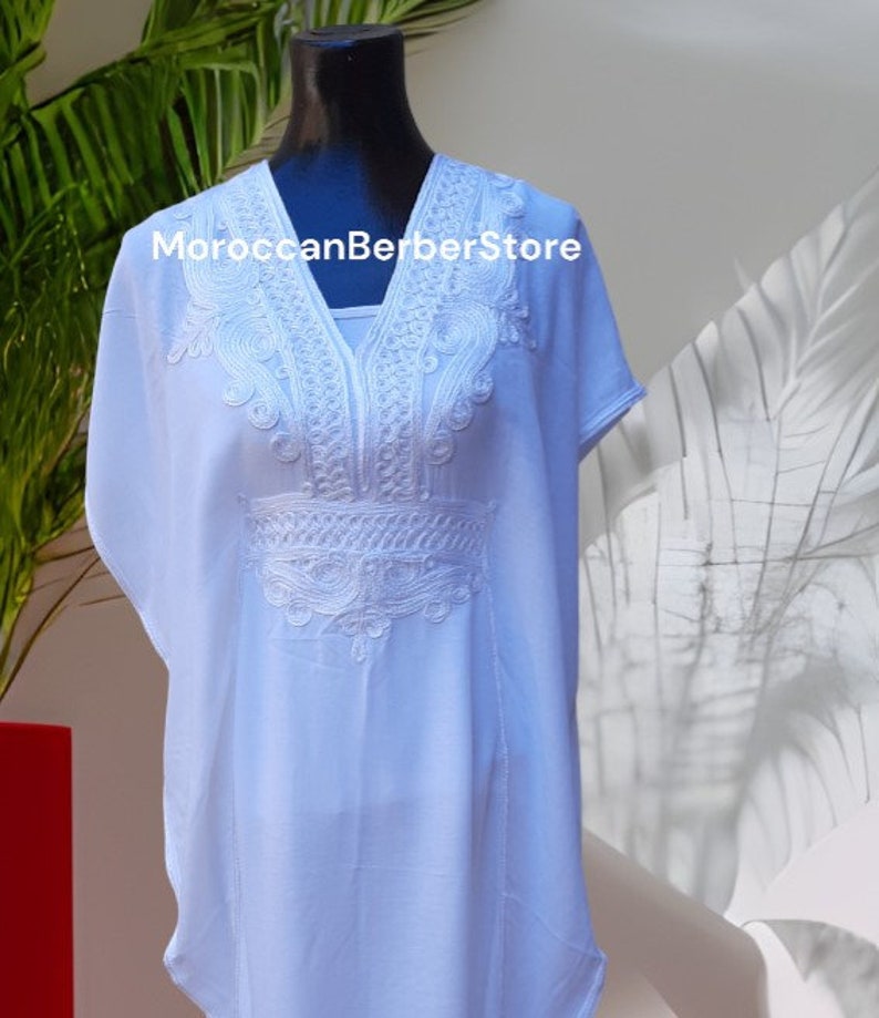 May include: A light blue kaftan with white embroidery. The embroidery features a geometric pattern around the neckline and a floral design on the chest. The text "MoroccanBerberStore" is visible on the garment.