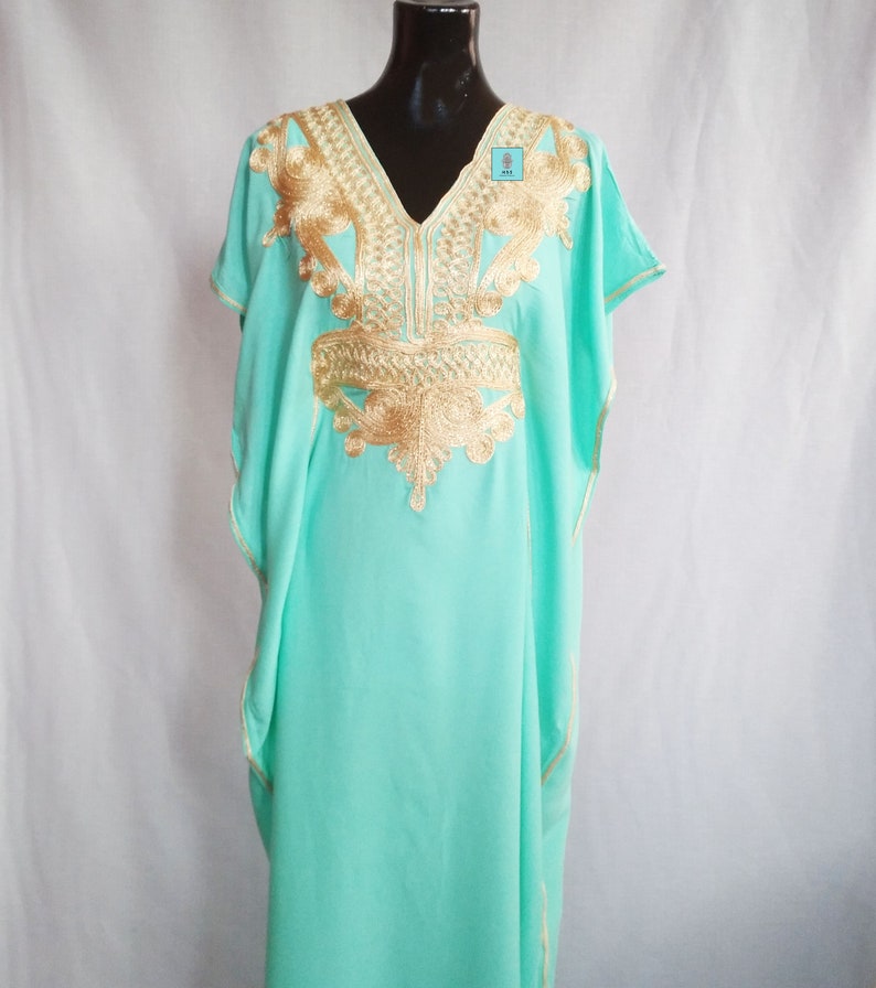 Puede incluir: Una kaftan verde menta con bordados dorados. La kaftan tiene cuello en V y mangas cortas. El bordado es intrincado y detallado, y cubre el escote, los hombros y el pecho de la prenda.