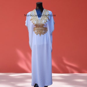 May include: A white kaftan with gold embroidery, featuring a V-neckline and short sleeves. The kaftan is displayed on a mannequin against a red background. The text "MoroccanBerberStore" is visible on the image.