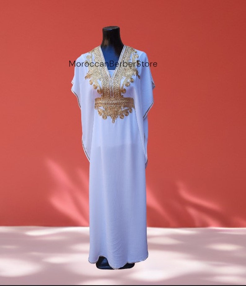 May include: A white kaftan with gold embroidery, featuring a V-neckline and short sleeves. The kaftan is displayed on a mannequin against a red background. The text "MoroccanBerberStore" is visible on the image.