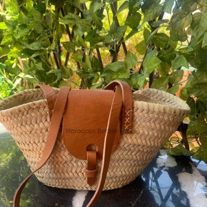 May include: Woven straw tote bag with a brown leather flap and shoulder strap. The bag has a wide opening and a natural, textured appearance. The leather flap is secured with a buckle. The text "Moroccan Berber Store" is visible on the bag.