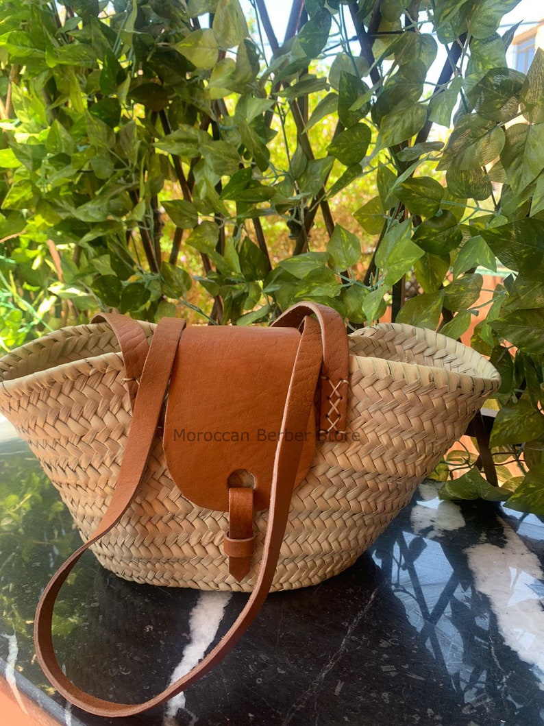 May include: Woven straw tote bag with a brown leather flap and shoulder strap. The bag has a wide opening and a natural, textured appearance. The leather flap is secured with a buckle. The text "Moroccan Berber Store" is visible on the bag.
