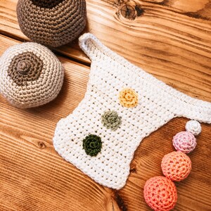Op de afbeelding: Gehaakte items op een houten oppervlak. De afbeelding toont twee ronde, tepelvormige gehaakte stukken, een wit gehaakt item met drie bloemvormige versieringen en een reeks gehaakte ballen in verschillende kleuren.