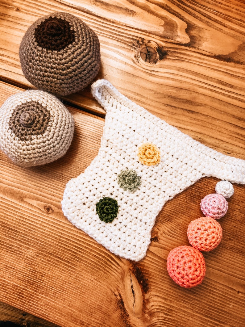 Op de afbeelding: Gehaakte items op een houten oppervlak. De afbeelding toont twee ronde, tepelvormige gehaakte stukken, een wit gehaakt item met drie bloemvormige versieringen en een reeks gehaakte ballen in verschillende kleuren.