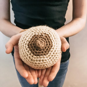 Op de afbeelding: Een rond, gehaakt object met een lichtbeige basis en een donkerder bruin cirkelvormig midden met een kleine, verhoogde, puntige top in het midden. Het item is gemaakt met zichtbare garensteken, wat zorgt voor een getextureerd, bolvormig en zacht uiterlijk.