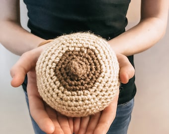 May include: A round, crocheted object featuring a light beige base and a darker brown circular center with a small, raised, pointed tip in the middle. The item is crafted with visible yarn stitches, creating a textured, spherical, and soft appearance.