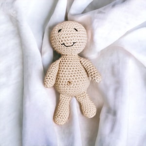 May include: A crocheted doll with a smiling face, beige in color, lying on a white fabric background.