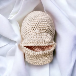 Puede incluir: Un gorro de bebé beige tejido a ganchillo con un borde doblado y un pequeño diseño texturizado en la parte superior. El interior del borde es de color rosa claro. El gorro se muestra sobre una tela blanca y sedosa.