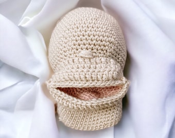 May include: A crocheted beige baby hat with a folded brim and a small, textured design on the top. The interior of the brim is a light pink color. The hat is displayed on a white, silky fabric.