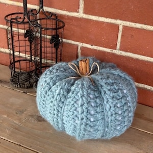 Puede incluir: Una calabaza tejida de color azul claro con un tallo de canela y un lazo de cuerda. La calabaza está sobre una superficie de madera frente a una pared de ladrillo. Una cesta de metal negro está en el fondo.
