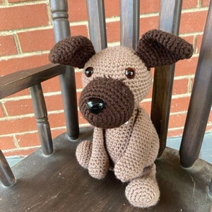 May include: A crocheted brown and tan dog toy sitting on a wooden chair. The dog has brown ears and a brown nose.