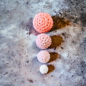 Op de afbeelding: Vier gehaakte bollen in een verticale lijn, elk in een andere maat en tint roze. De grootste is koraal van kleur, gevolgd door een medium roze, een lichtroze en een kleine witte bol. De bollen zijn gerangschikt op een gevlekte grijze ondergrond.