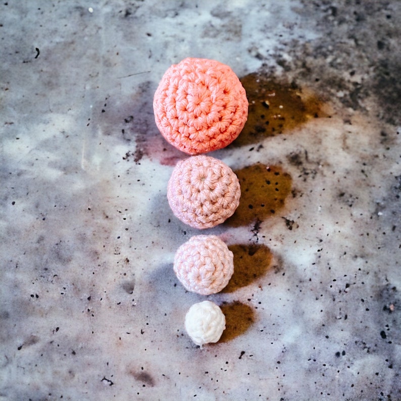 Op de afbeelding: Vier gehaakte bollen in een verticale lijn, elk in een andere maat en tint roze. De grootste is koraal van kleur, gevolgd door een medium roze, een lichtroze en een kleine witte bol. De bollen zijn gerangschikt op een gevlekte grijze ondergrond.