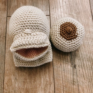 Op de afbeelding: Twee gehaakte items op een houten oppervlak. Eén is een lichtbeige wantvormig object met een roze binnenkant. De andere is een lichtbeige bal met een bruine cirkel in het midden. De items zijn handgemaakt.