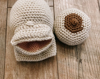 May include: Two crocheted items on a wooden surface. One is a light beige mitten-shaped object with a pink interior. The other is a light beige ball with a brown circle in the center. The items are handmade.