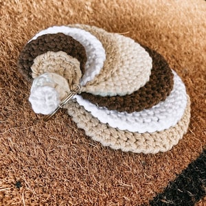 Puede incluir: Un conjunto de cinco círculos de ganchillo en tonos marrones, beige y blancos. Los círculos están apilados uno encima del otro, creando un efecto de capas.