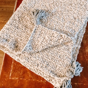 Puede incluir: Una manta de felpa blanca y gris con flecos de borla grises en los extremos.