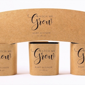 May include: Kraft paper baby shower favors with the text "Watch Me Grow" in elegant script. The favors include the baby's name and date. The design features a curved paper sleeve and three cylindrical containers.