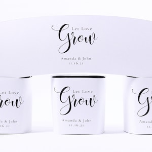 May include: White wedding favors with the text "Let Love Grow" in elegant script. The names "Amanda & John" and the date "11.16.21" are also printed on the favors. The favors are cylindrical and have a black interior.
