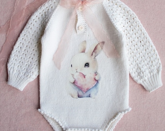 May include: White knitted baby outfit with a bunny design and a matching bonnet. The outfit has a pink ribbon and buttons.