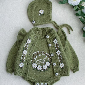 Könnte beinhalten: Olivgrünes Strick-Baby-Outfit-Set, bestehend aus einem Strampler und einer Mütze. Der Strampler hat weiße Gänseblümchen-Stickereien und einen Blumenkranz mit dem Buchstaben "A". Die Mütze hat eine Gänseblümchen-Akzent. Weiße Knöpfe.