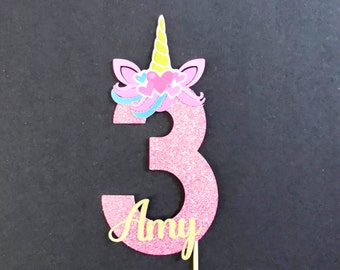 May include: Pink glitter cake topper with a unicorn head and the number 3. The unicorn has a yellow horn, pink ears, and blue and pink mane. The name Amy is written in gold glitter below the number.
