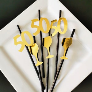 Può includere: Sei cannucce nere con ritagli in lamina d'oro del numero 50 e un bicchiere di vino. Le cannucce sono disposte su un piatto bianco.