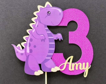 May include: Purple dinosaur cake topper with a pink glitter number 3 and the name "Amy" written in gold glitter.