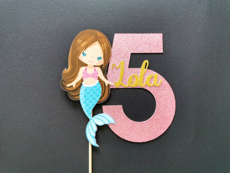 Puede incluir: Topper de pastel de n&uacute;mero cinco de purpurina rosa con un personaje de sirena y el nombre "Lola" en purpurina dorada.